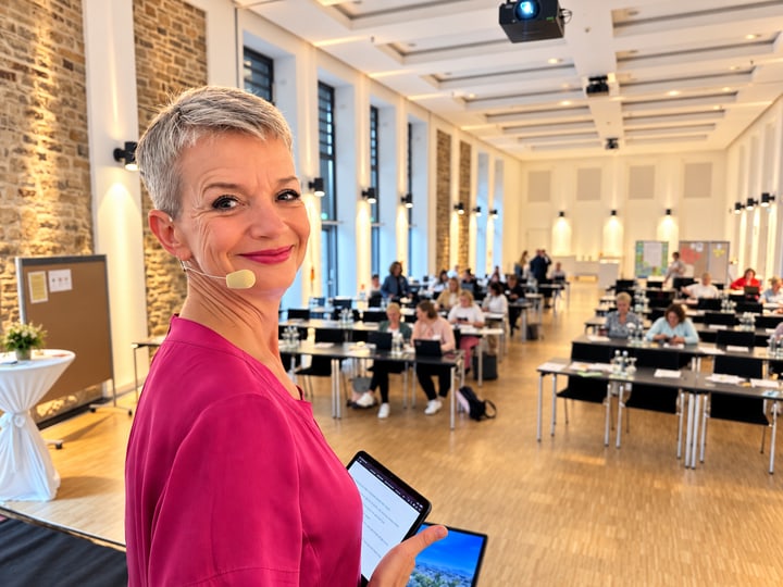 Andrea Kaden mit Tablet vor einem Konferenzraum mit Teilnehmenden